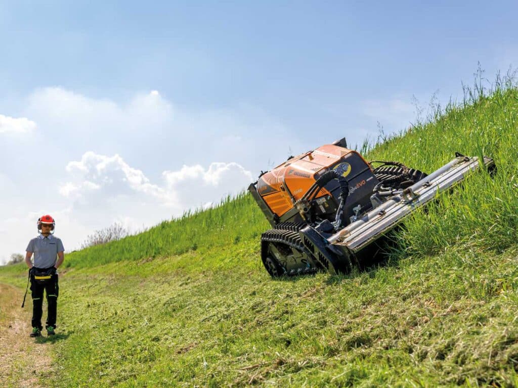 Remote-controlled mowing machine operating on a grassy hillside under blue sky.
