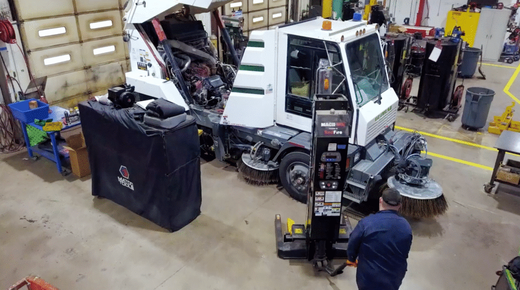 Mechanic performing heavy equipment maintenance on a street sweeper in a workshop.