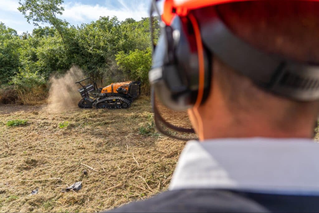 Operator oversees robotic mower clearing land in a rural environment.
