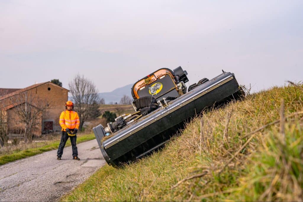Man operating a slope mower on a rural road in a scenic landscape.