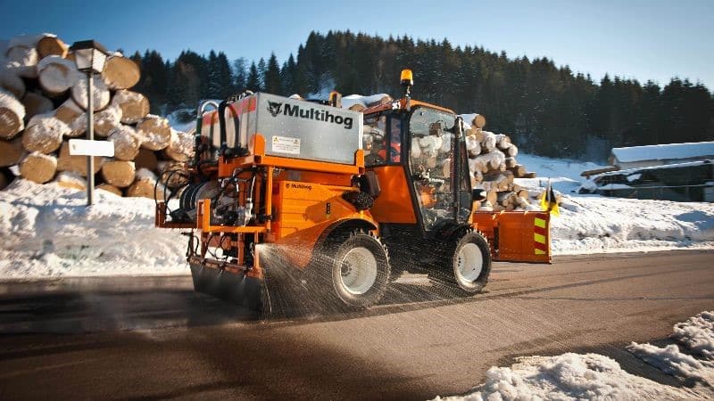 Multihog vehicle clearing snow while surrounded by logs in a winter landscape.