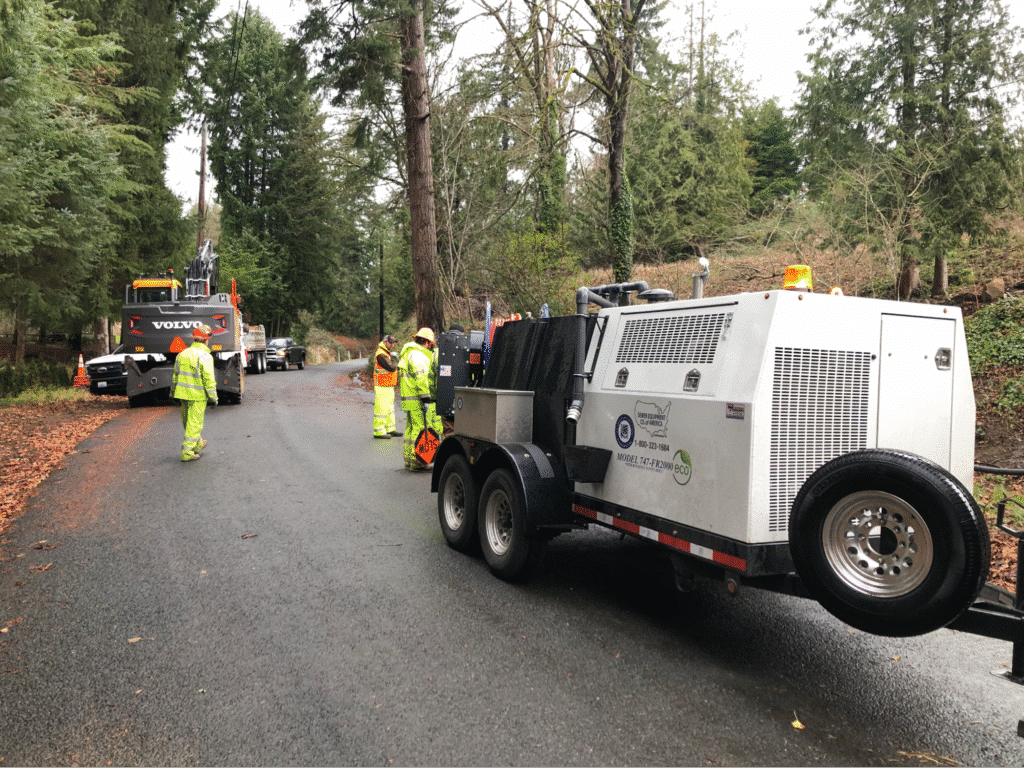 Workers in safety gear manage sewer jetter equipment in a forested area.