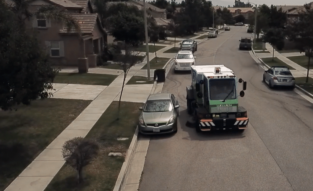 Street cleaning vehicle on residential street while parked cars line the curb.