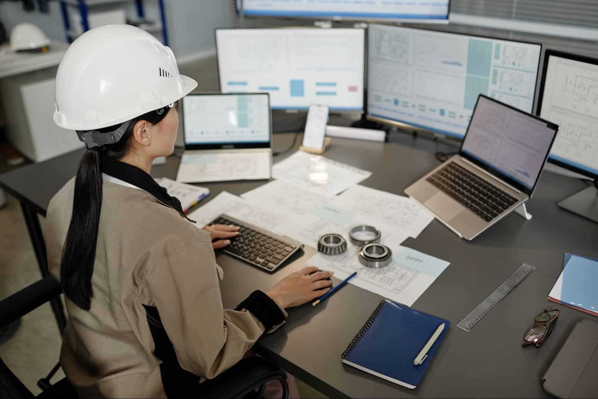 Engineer working at a desk with multiple monitors, design plans, and safety gear.