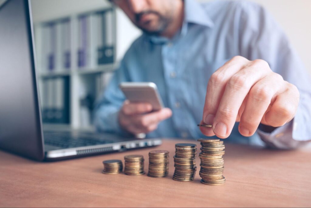 Man managing finances with coins and smartphone near a laptop.
