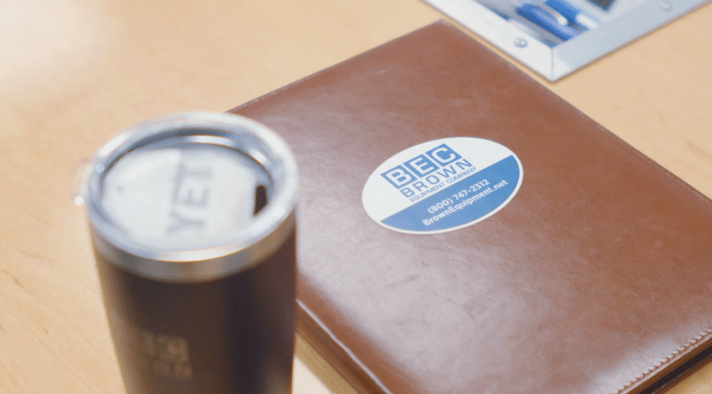 Coffee cup beside a BEC branded notebook on a wooden table.