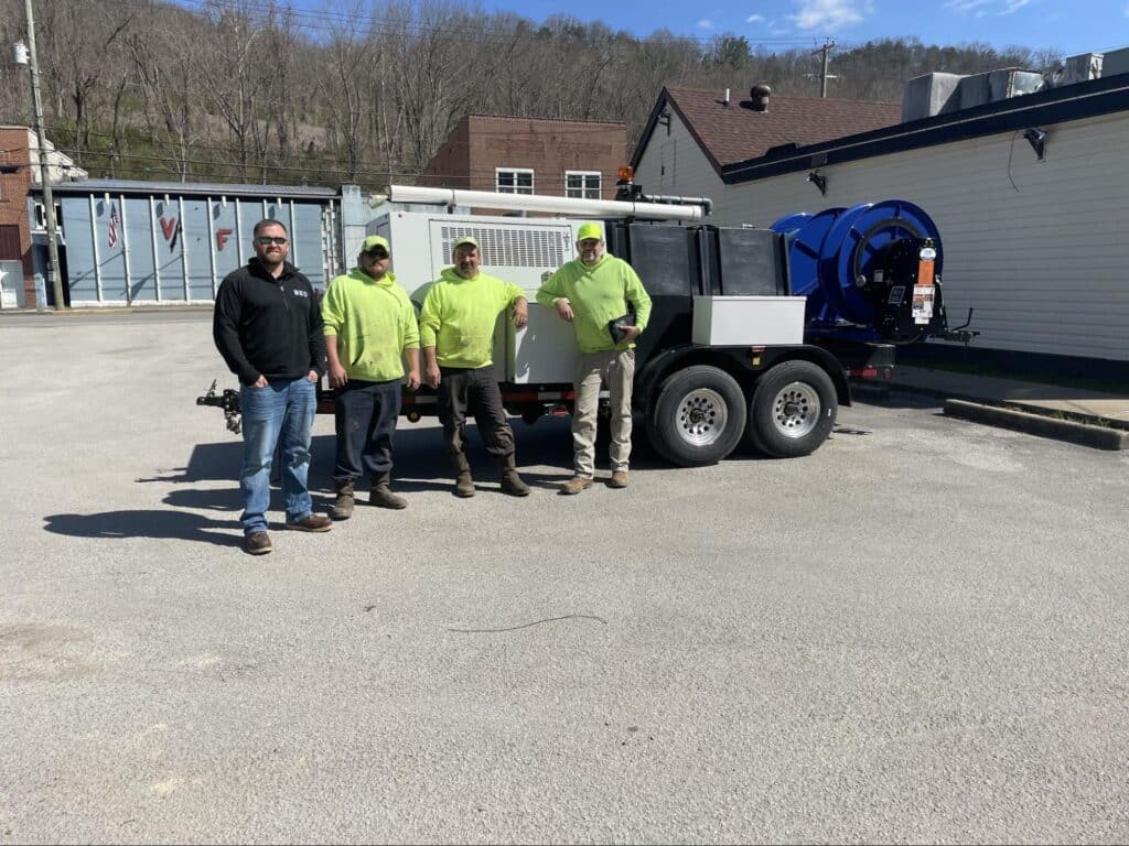 Four workers in bright shirts stand by jetting equipment in a parking lot.