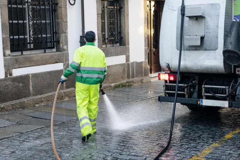 Worker cleaning cobblestone streets with a truck-mounted water hose.