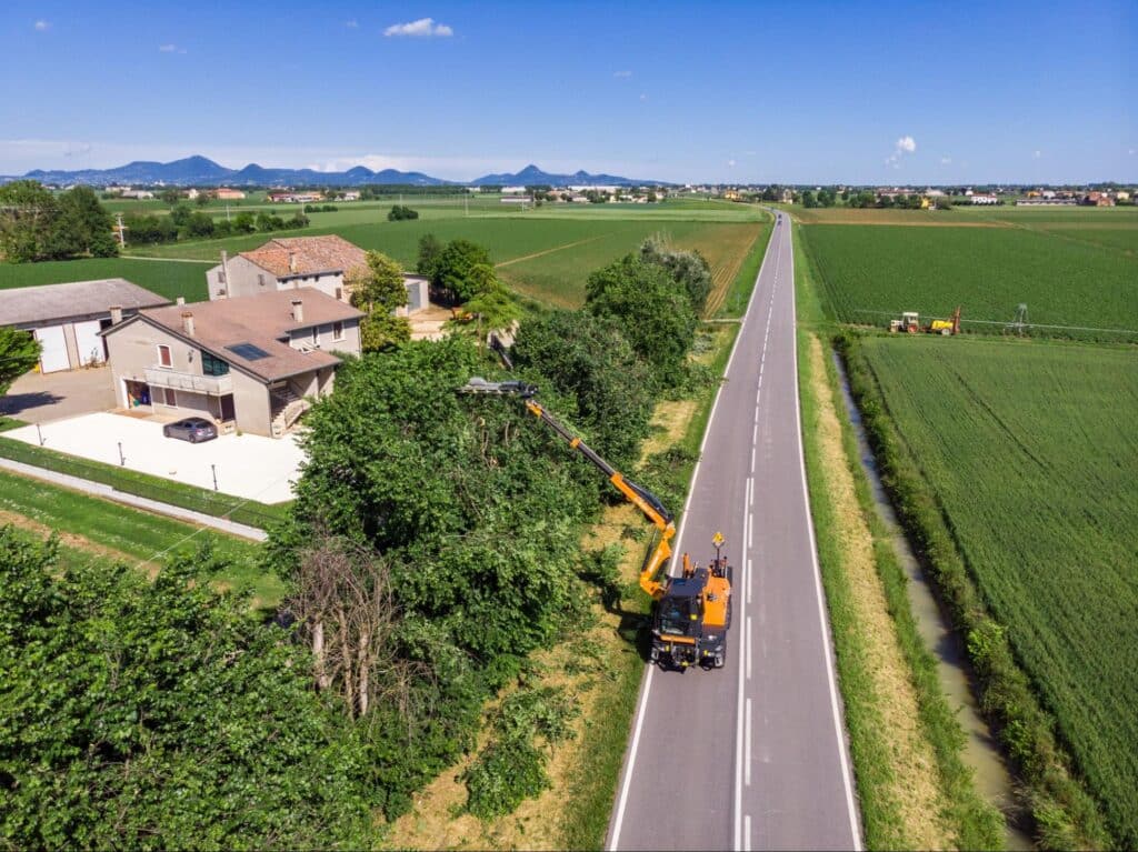 Tree trimming boom attachment on equipment on a rural road near fields and mountains.