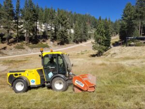 Yellow Multihog mower clearing grass in a picturesque forest landscape under blue skies.