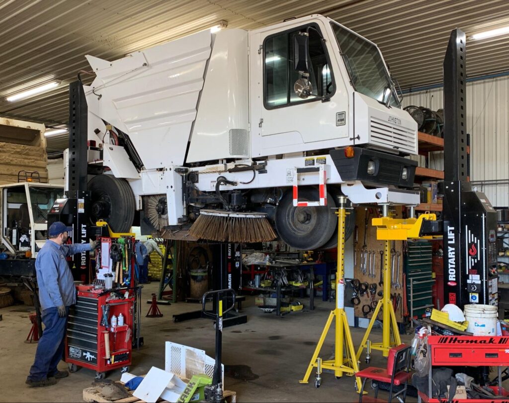Street sweeper undergoing maintenance in a repair shop with tools and equipment.