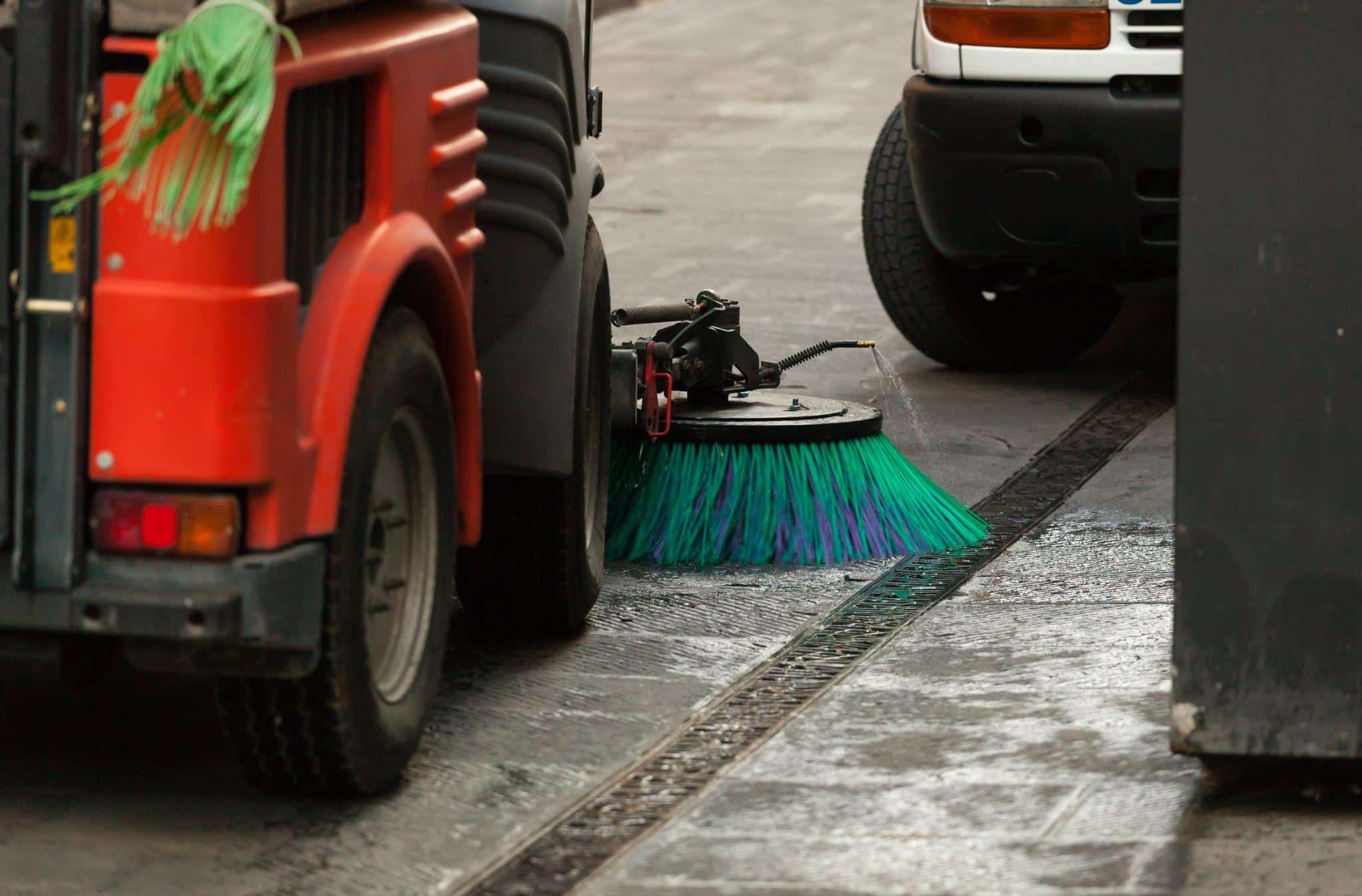 Urban street sweeper cleaning pavement alongside parked cars.