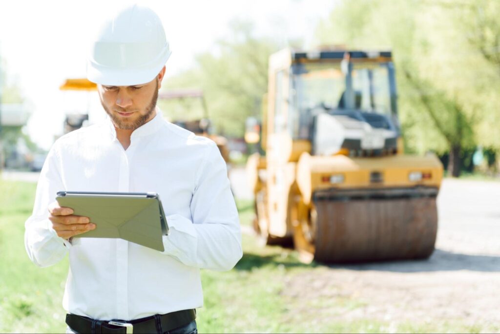 Construction manager operating a tablet while overseeing machinery at a job site.