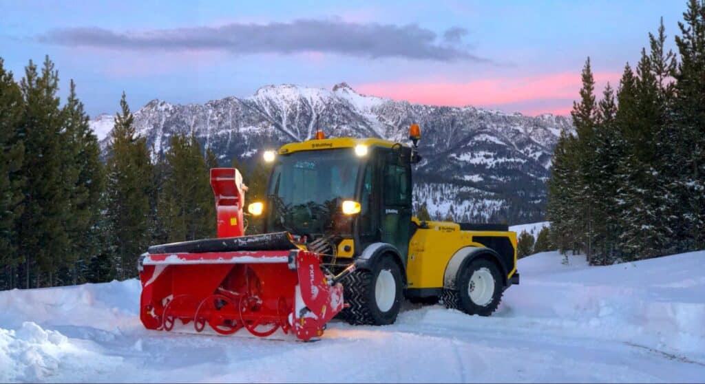 Snow plow clearing snowy mountain roads at sunset in winter scenery.