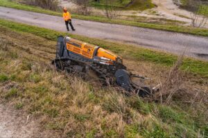 Energreen Robo machine efficiently clears grass and brush on hillside near gravel road.